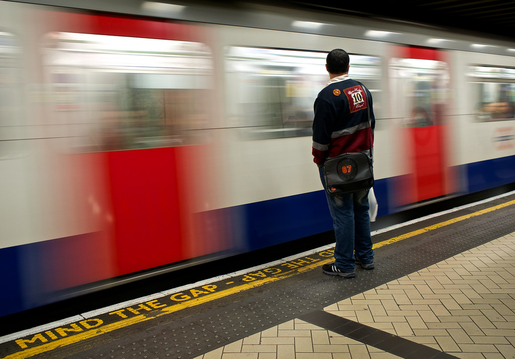 A red London bus passing a Tube station entrance with commuters on a busy street.