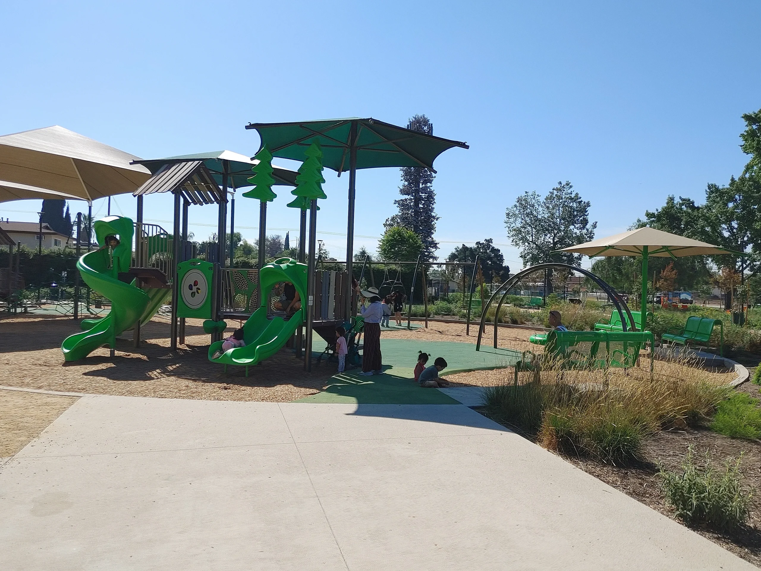Children playing on new equipment in a renovated Zamora Park, El Monte, with adults walking on paved paths