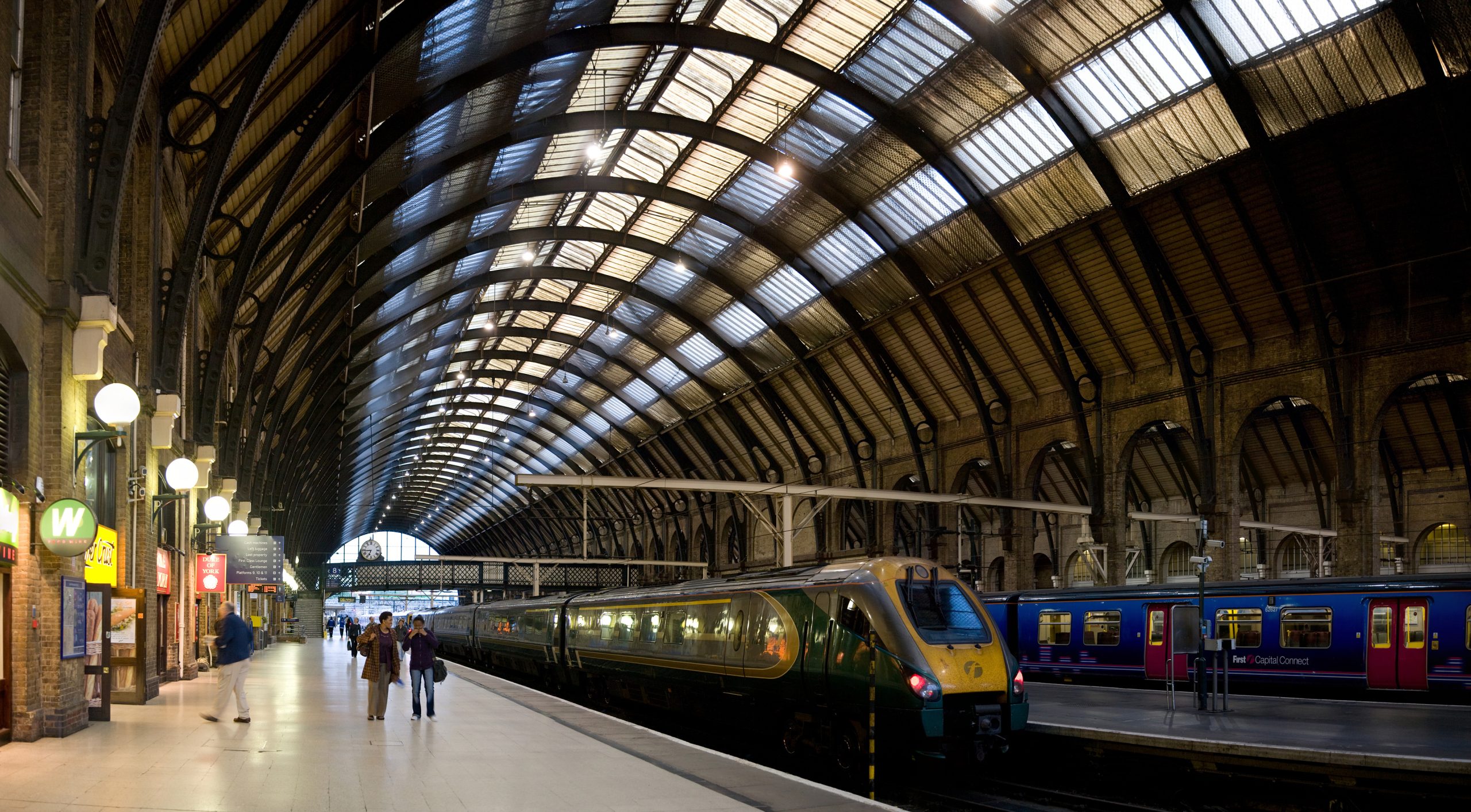 A busy London Underground station platform with a train arriving, passengers waiting, and a Transport for London roundel logo visible.