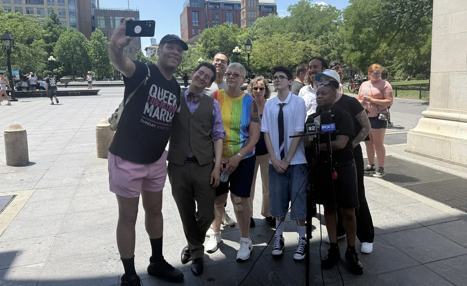 A diverse crowd gathers in Washington Square Park, with some holding Pride flags and banners, as activists speak at a press conference.