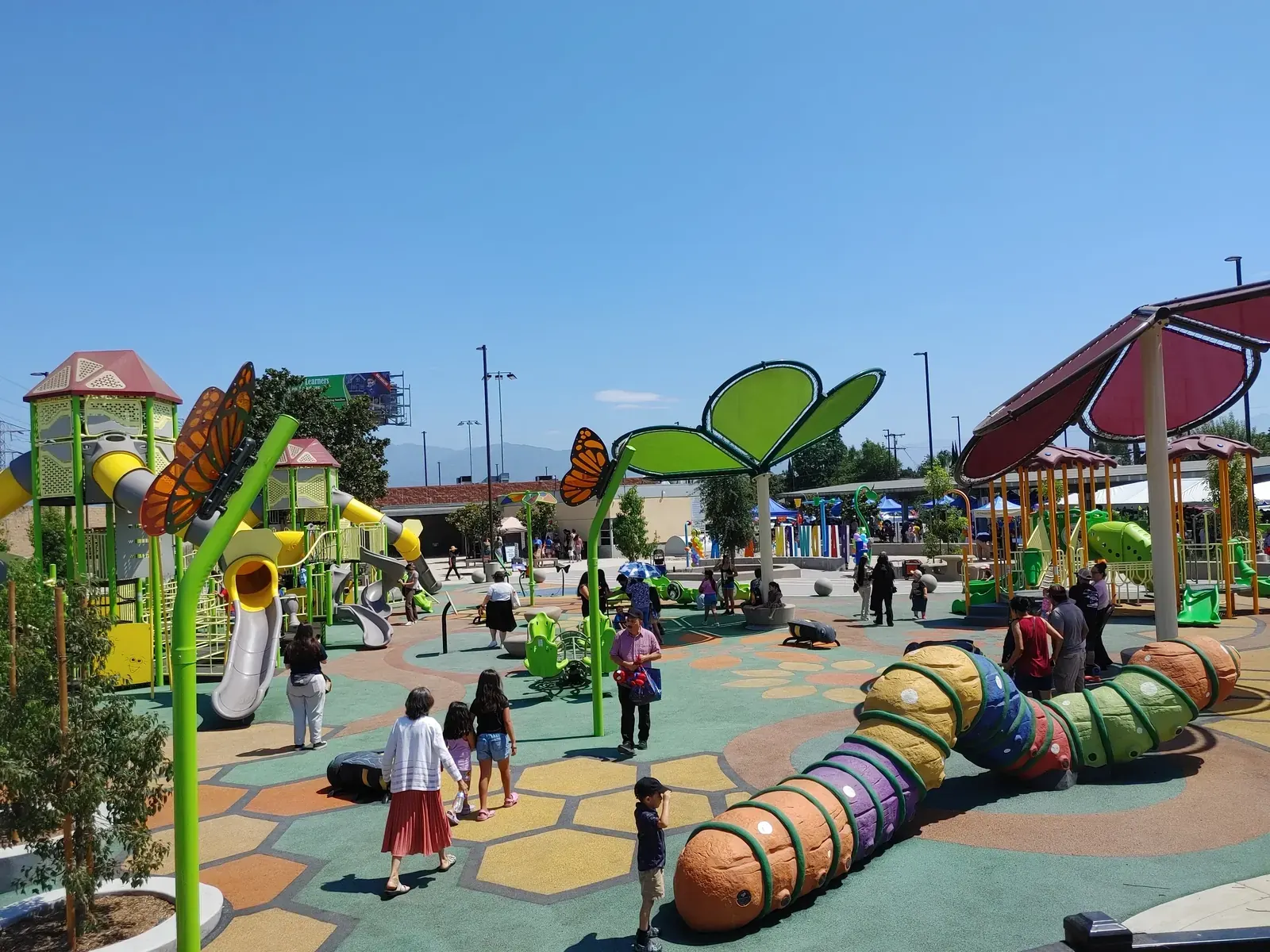 Children and adults enjoying an accessible, multi-generational playground in a redeveloped urban park, with new basketball courts and walking trails visible in the background.
