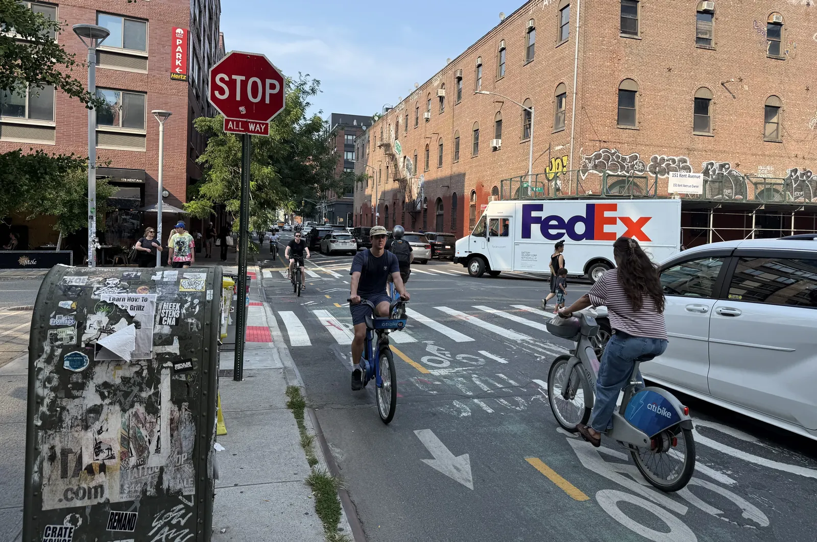 Cyclists and pedestrians on a narrow protected bike lane on Kent Avenue in Williamsburg, Brooklyn, with parked cars and a signalised intersection
